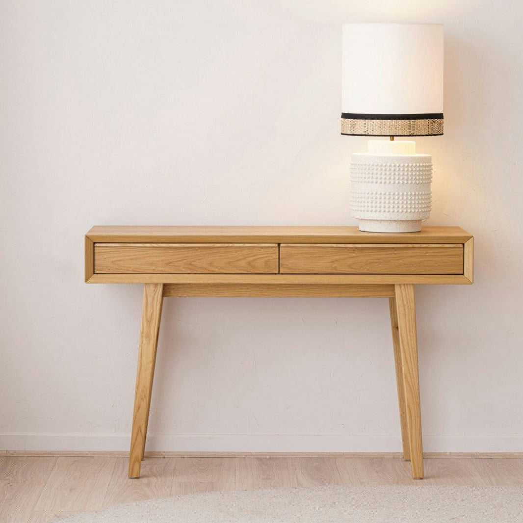 La Consola en Roble Escandinava Bergen, con dos cajones, se apoya contra una pared blanca y muestra una lámpara de cerámica blanca sobre su superficie.