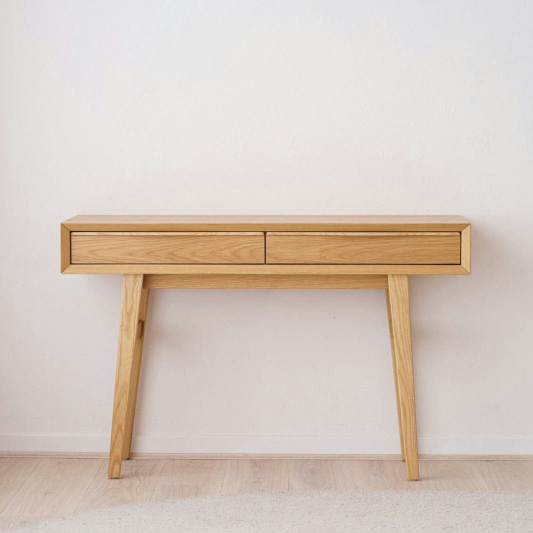 La Consola en Roble Escandinava Bergen con dos cajones y patas anguladas resalta sobre una pared blanca y suelo de madera clara.