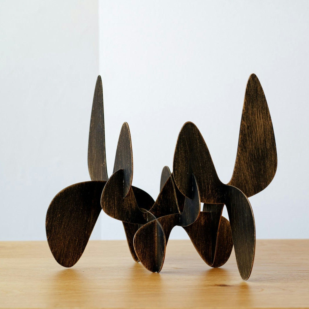 Figuras Decorativas en Metal Orgánico Lámina Oro presenta una escultura abstracta de madera con formas curvas entrelazadas y un toque de pan de oro en la superficie de madera, sobre un fondo blanco.