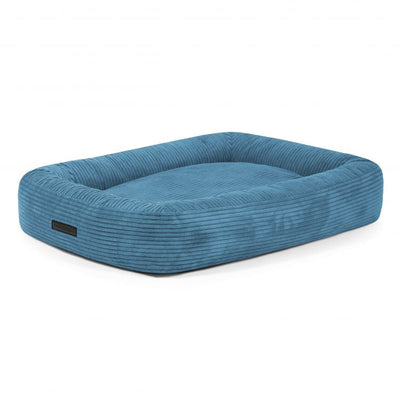 Cama para mascotas de pana azul rectangular con bordes elevados y una etiqueta negra en un lado, sobre un fondo blanco.