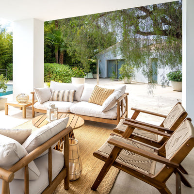 Patio exterior con muebles de madera acolchados, una mesa de centro redonda, una alfombra tejida y sillas, con vistas a una zona ajardinada y a una casa blanca con grandes ventanales.