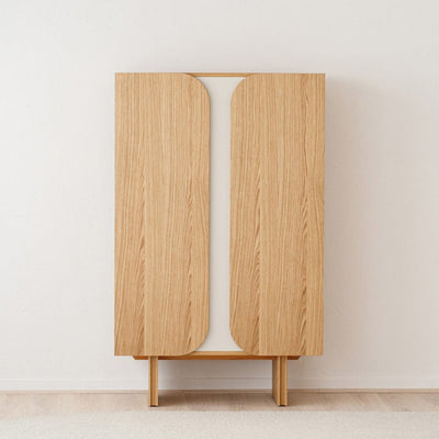 Un armario de madera con dos puertas verticales, colocado contra una pared blanca lisa, sobre un suelo de color claro.