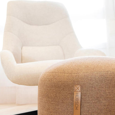 Un sillón tapizado en beige claro se coloca detrás de una otomana marrón con la etiqueta "HUBSCH" en una habitación luminosa y minimalista.