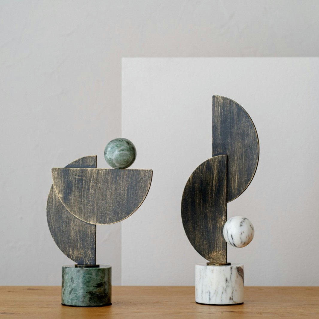 Dos esculturas abstractas descansan sobre una superficie de madera, con formas geométricas y bases de mármol verde y blanco, similares a las Figuras decorativas en mármol y metal Arco N1. Rematadas con elementos de madera y piedra, son perfectas para los entusiastas de la decoración de interiores que buscan detalles únicos.