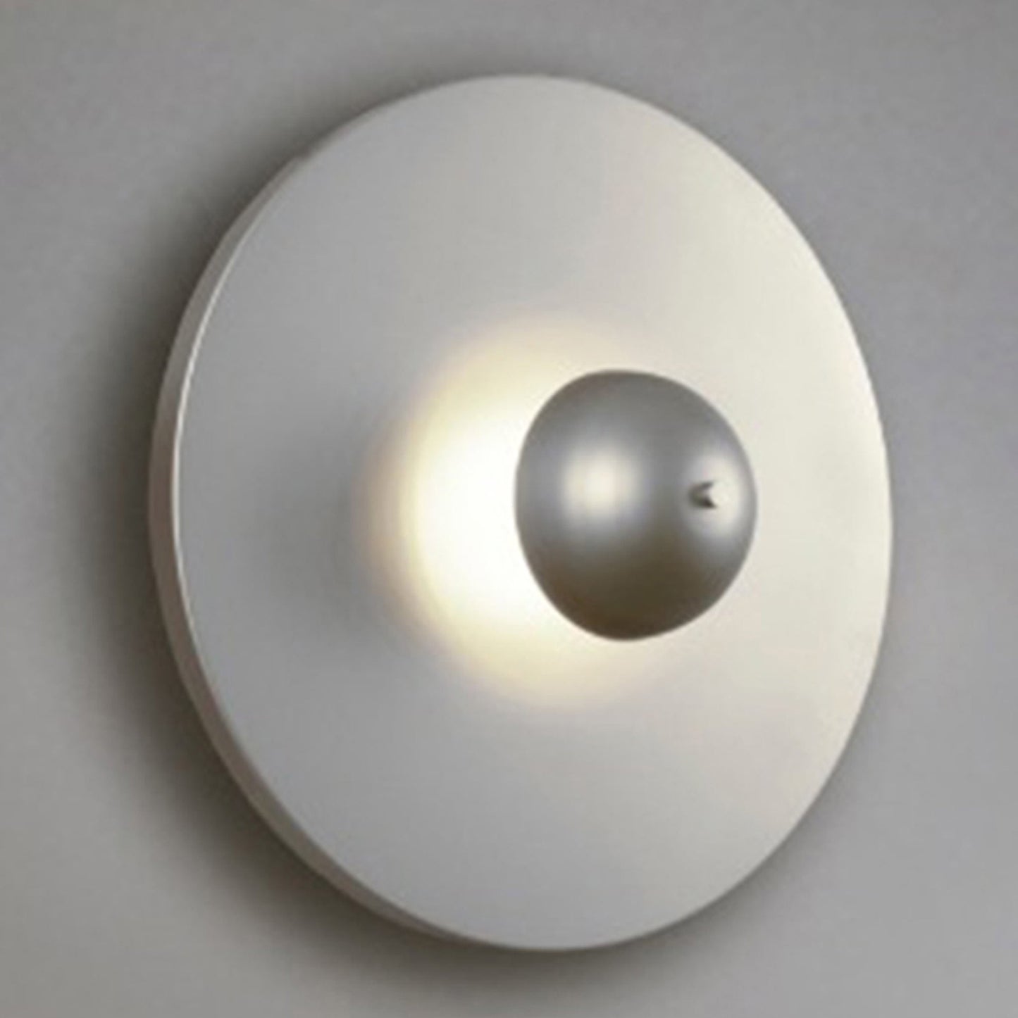 El aplique de pared en acero Disc presenta un diseño elegante con una esfera metálica central que emite un brillo suave en su superficie redonda plana y de color claro, ejemplificando la iluminación moderna.
