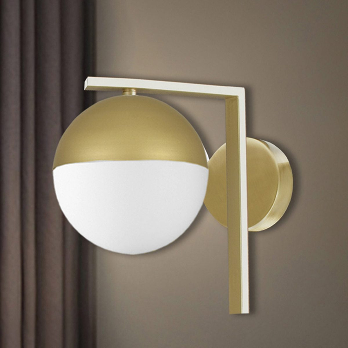 Aplique de pared en acero Janus con lámpara esférica blanca y dorada sobre soporte vertical dorado, montado en una pared beige, complementado por cortinas oscuras.