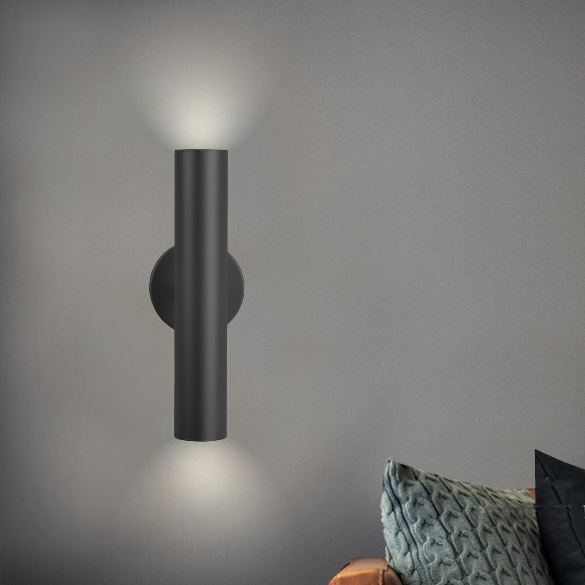 Aplique de pared de acero Met con iluminación hacia arriba y abajo en una pared gris, junto a un cojín estampado en el reposabrazos.