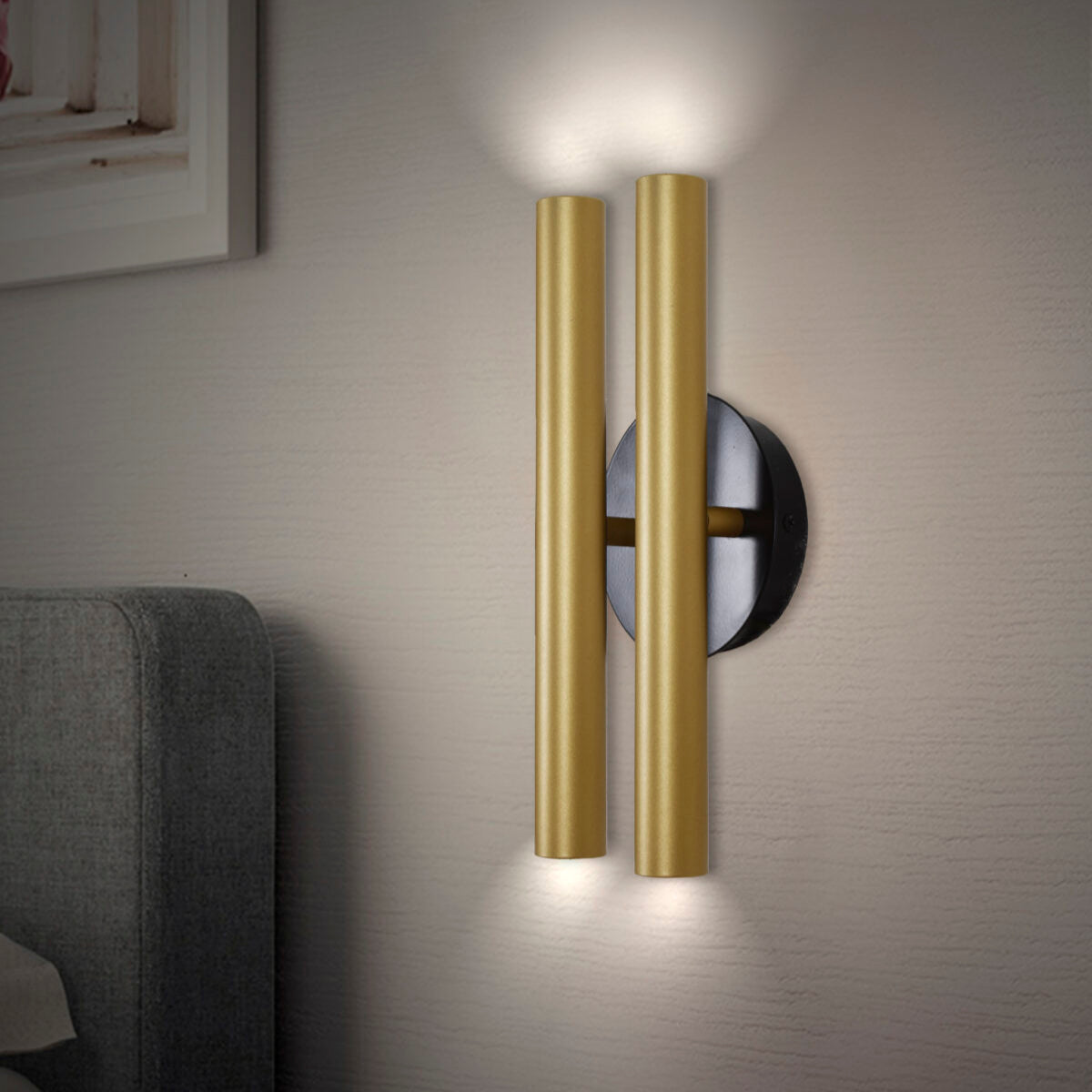 El aplique de pared en acero con 4 puntos de luz Caia presenta un diseño moderno, con tubos dorados y base negra, montado en una pared beige que emite una iluminación óptima desde ambos extremos.