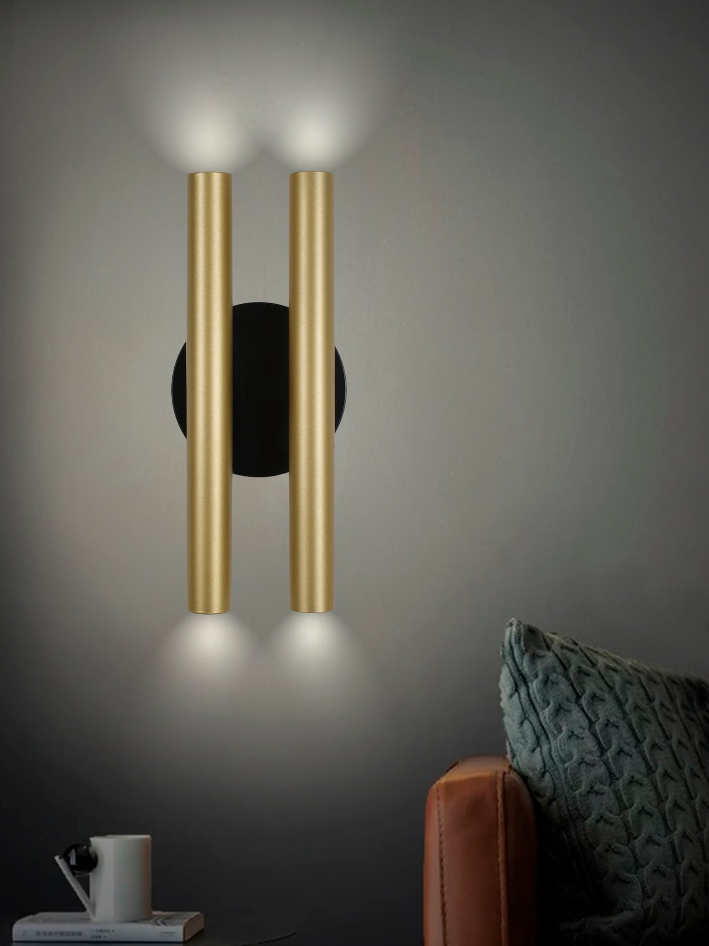 El Aplique de pared en acero con 4 puntos de luz Caia ofrece una iluminación perfecta con sus dos tubos verticales dorados. Cerca, una almohada con textura verde adorna el sofá marrón junto a una mesa pequeña con una taza de café y un libro, creando un ambiente de diseño moderno.