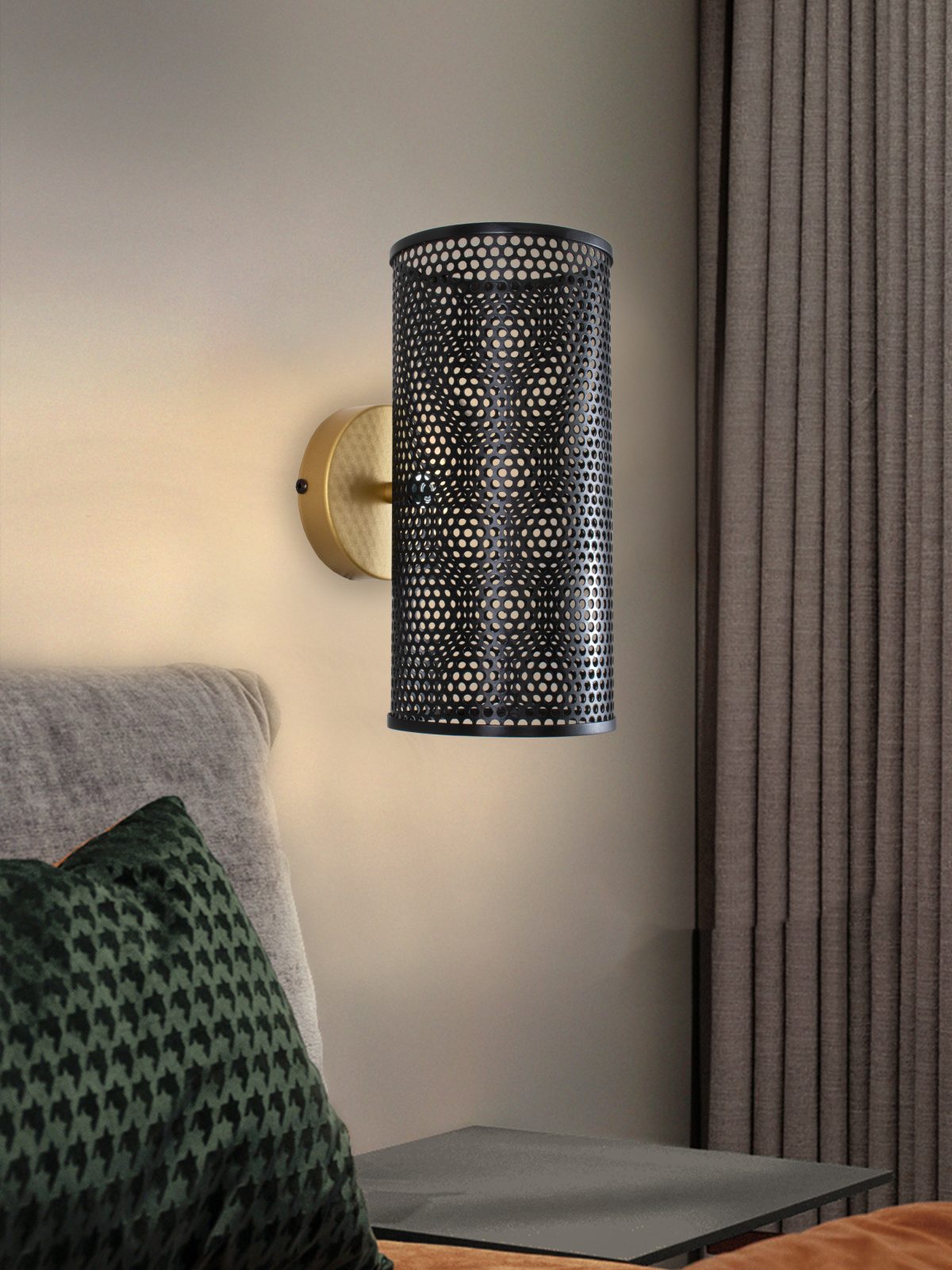 Un aplique de pared moderno con pantalla cilíndrica perforada ilumina la pared de un dormitorio y resalta su diseño moderno. Las cortinas cercanas complementan la almohada estampada verde de la cama, mientras que el aplique de pared en acero Mira en negro-oro agrega un toque elegante.