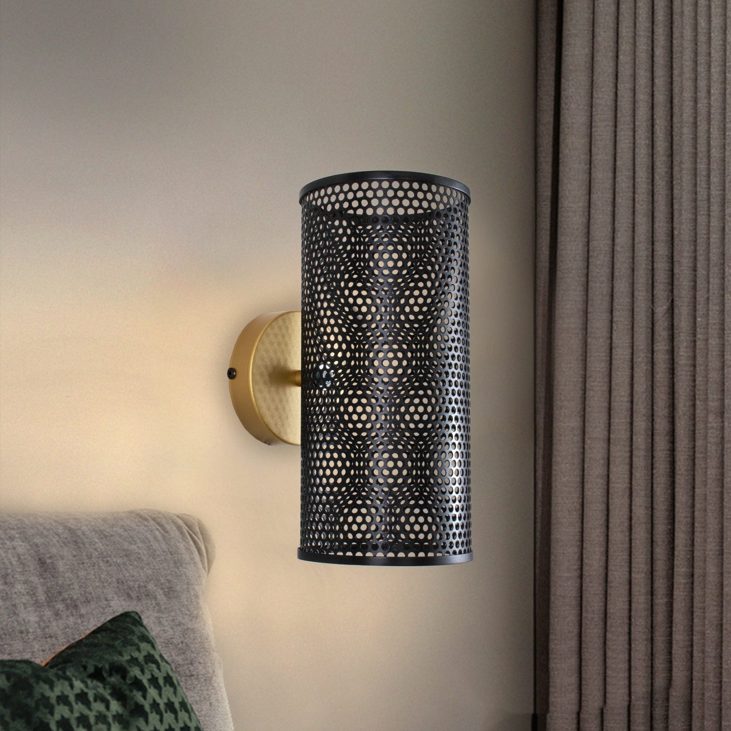 Una moderna lámpara de pared de acero Mira con pantalla cilíndrica negra perforada y base de latón en un elegante acabado acero negro-oro, montada junto a un cabecero gris y cortinas beige.