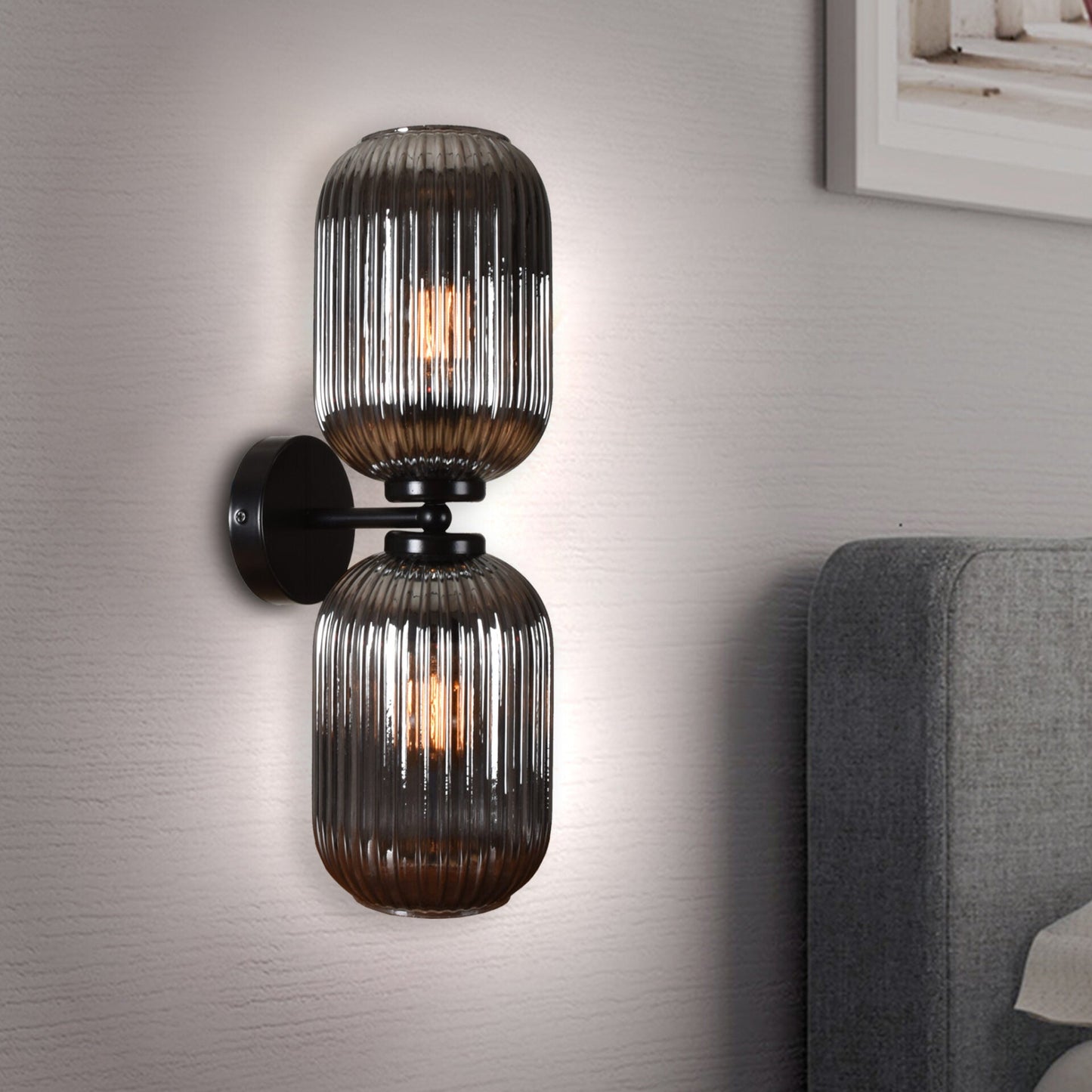 El Aplique de pared en acero y cristal Kairo, un aplique de pared de doble cristal con diseño acanalado, proyecta luz difusa sobre una pared gris al lado del sofá, realzándola con iluminación moderna.