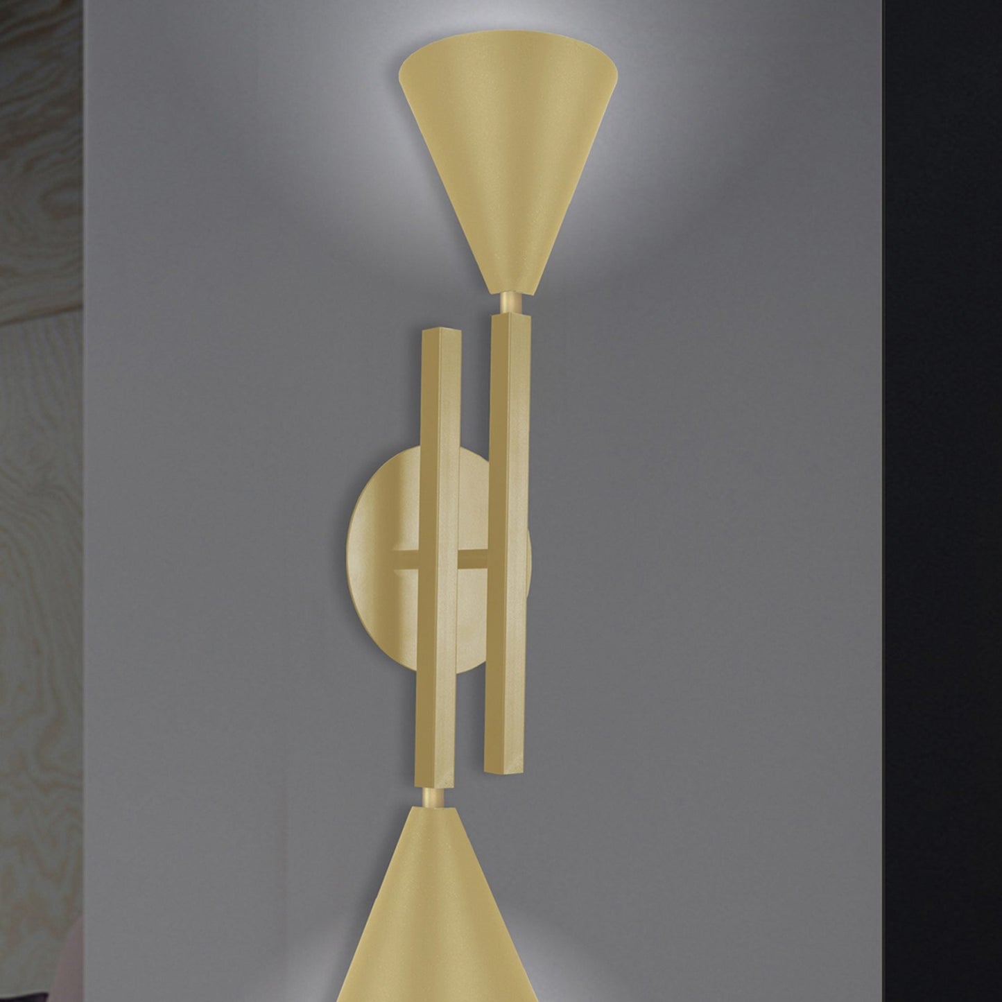 Aplique de pared moderno dorado con dos luces en forma de cono orientadas en direcciones opuestas sobre una pared gris. Esta pieza de iluminación moderna industrial, similar al Aplique de pared en acero con 2 puntos de luz Big Nyx, combina sin esfuerzo funcionalidad y estilo.