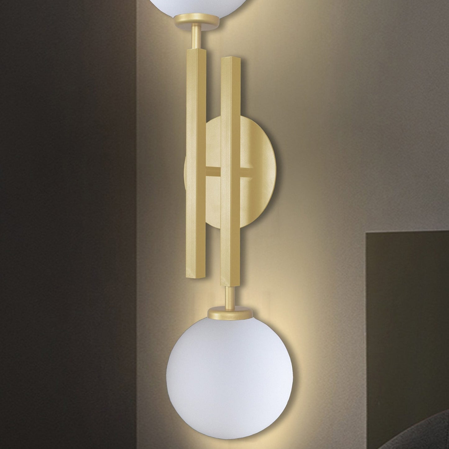 El aplique de pared de acero con 2 puntos de luz Nysa, con un diseño moderno de aplique de pared con acabado dorado y dos globos de vidrio esmerilado alineados verticalmente contra una pared beige, agrega sofisticación a cualquier habitación.