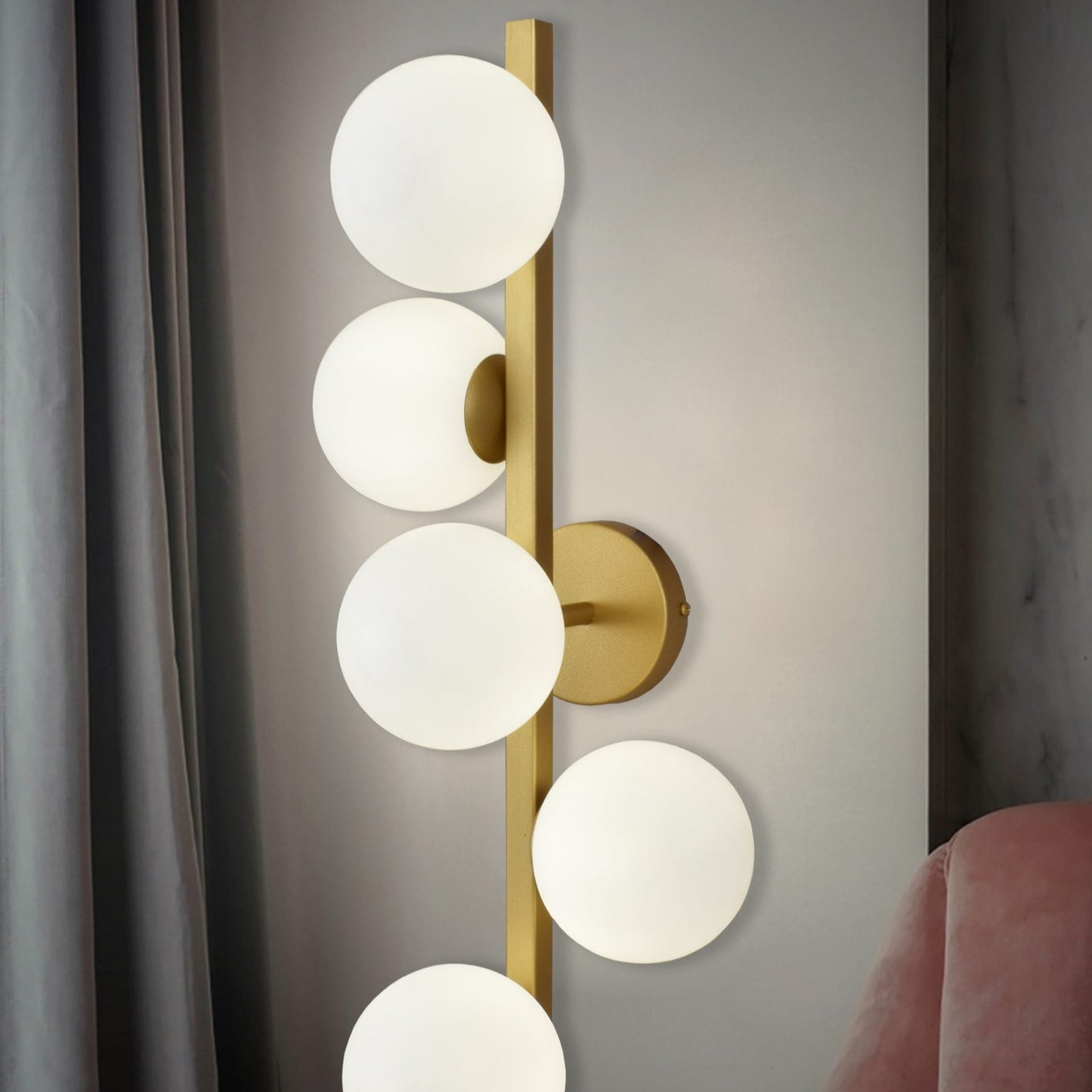 El aplique de pared de acero Holly, una elegante lámpara de pared con una varilla dorada vertical y cinco bombillas esféricas blancas, añade un diseño contemporáneo junto a la silla tapizada en rosa y la cortina gris.