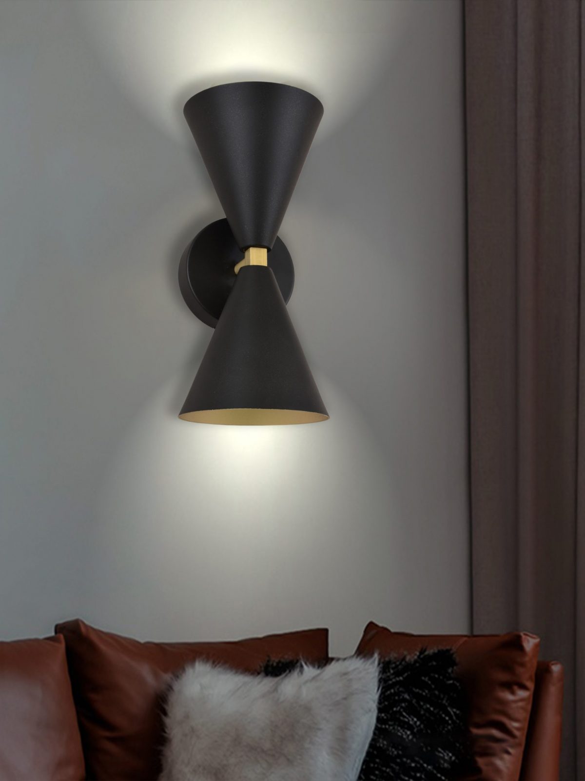 El aplique de pared en acero con 2 puntos de luz Nyx, en forma de cono negro mate, emite un brillo suave, montado elegantemente sobre un sofá de cuero marrón con una almohada en blanco y negro, infundiendo a la habitación un diseño contemporáneo.