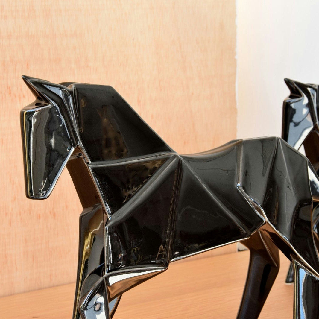 Dos esculturas de cerámica negra brillante de Figuras decorativas en cerámica Negro brillo Caballo Origami, que se asemejan a caballos angulares y estilizados, se asientan sobre una superficie de madera contra una pared de color claro, abrazando la estética contemporánea.