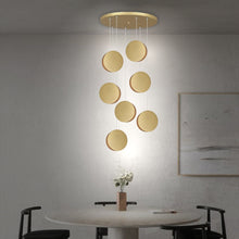  Un comedor moderno presenta una mesa redonda, cuatro sillas negras y la Lámpara de techo Colgante Moderna Dorada 3223-7C-3 con elementos circulares dorados. El espacio está adornado con una pintura minimalista y un pequeño jarrón de flores, todo bajo una elegante iluminación interior.