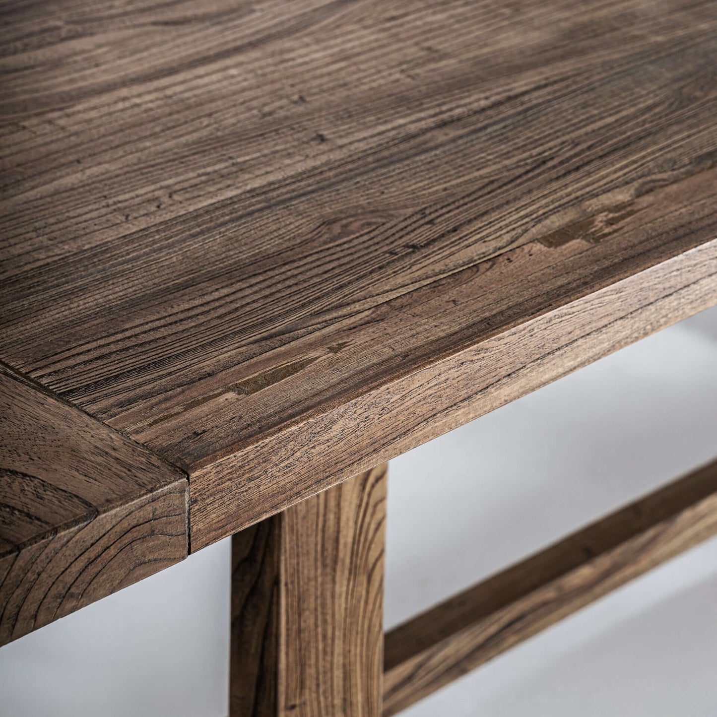 Detalle de la mesa de comedor NORVIK, de madera de olmo marrón (220×100×75 cm), que muestra su veteado natural y sus robustas patas sobre un fondo claro.