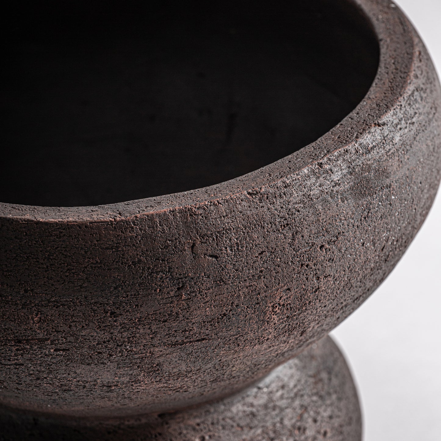 Detalle del Jarrón en terracota mate 26×26×26 cm - contemporáneo - OKAR P, con superficie marrón oscuro texturada, borde ancho y cuerpo redondeado sobre fondo claro liso.