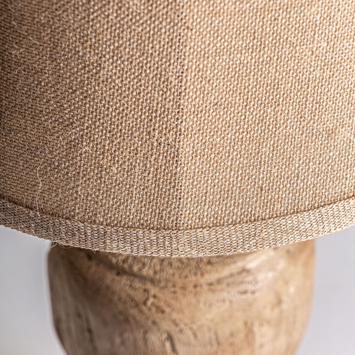 Primer plano de una pantalla de tela beige con un patrón texturizado, similar a una pantalla de yute, que reposa sobre una base de madera intrincadamente tallada con un acabado marrón envejecido, que aparece en la Lámpara de sobremesa en resina marrón Panerra.