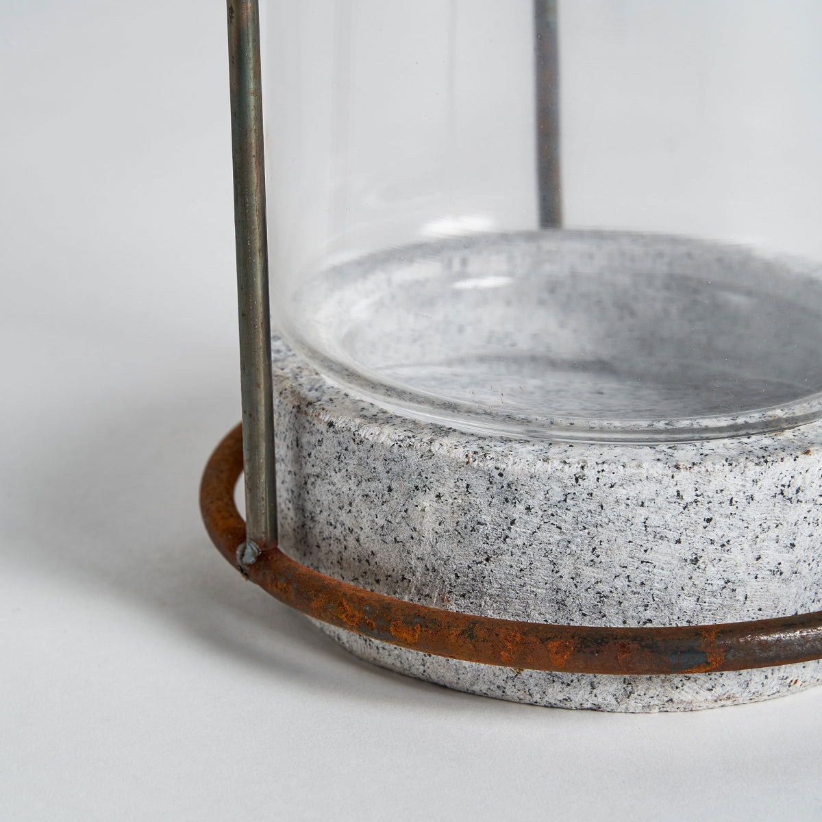 El Portavela en vidrio y piedra Lumina presenta un recipiente cilíndrico de vidrio con estructura de metal y base de piedra, resaltando la calidez y elegancia del diseño contemporáneo con un anillo de metal rústico en la base.