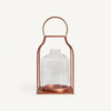 El Faro de vidrio y acero cobre es un recipiente de vidrio con marco metálico color cobre y asa superior, similar a una linterna industrial. Ideal para crear efectos de iluminación cálidos, se presenta sobre un fondo blanco.