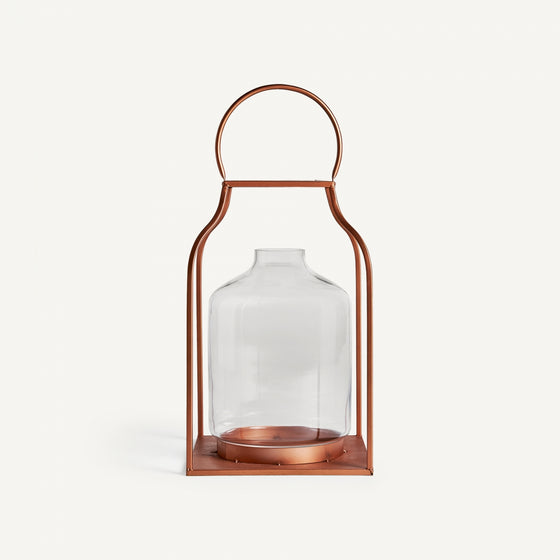 El Faro de vidrio y acero cobre es un recipiente de vidrio con marco metálico color cobre y asa superior, similar a una linterna industrial. Ideal para crear efectos de iluminación cálidos, se presenta sobre un fondo blanco.
