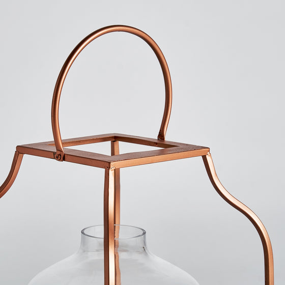 El Farol en vidrio y acero cobre Faro es una linterna de metal de color cobre con parte superior cuadrada y asa arqueada, vidrio transparente en la parte inferior y acabado industrial, ideal para agregar una iluminación cálida a cualquier espacio.