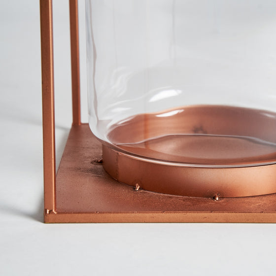 El Farol en vidrio y acero cobre Faro cuenta con un recipiente de vidrio transparente con una base circular sobre un marco de metal cuadrado de color cobre, perfecto para agregar una iluminación cálida y un estilo industrial a cualquier espacio.