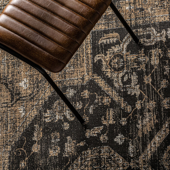 Una silla de cuero acanalado de color marrón con patas de metal negro reposa sobre la clásica alfombra de viscosa Keisi (340x240 cm), que presenta un patrón vintage texturizado en tonos beige y oscuros.