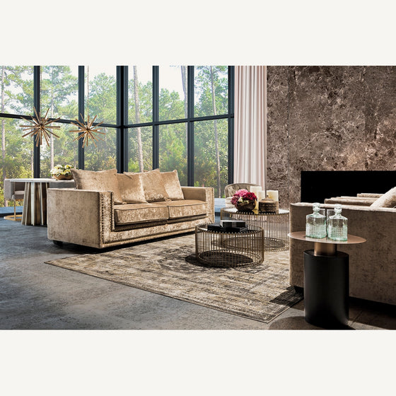 Moderna sala de estar con sofás beige, la clásica alfombra en viscosa Keisi (340x240 cm), mesa de centro redonda con elementos de decoración, grandes ventanales, pared de acento de piedra y un área de comedor y vegetación en el fondo que le agrega un toque vintage.