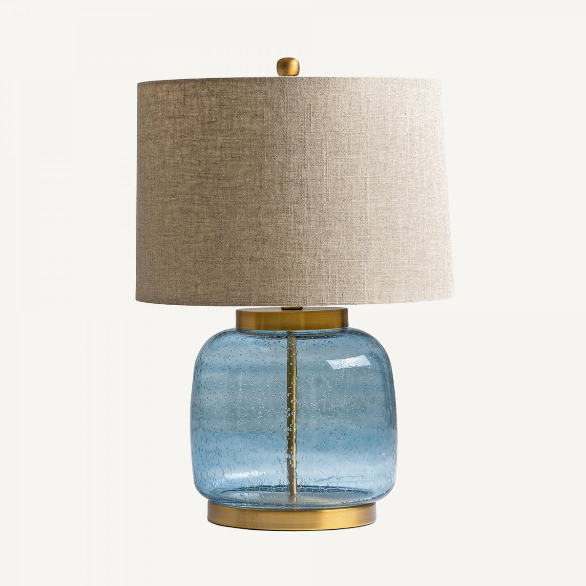 La Lámpara de mesa en vidrio azul y detalles dorados Bluey, con pantalla de tela beige y base de vidrio azul art deco, acentos estilo dorados y luz cálida, resalta sobre un fondo blanco.