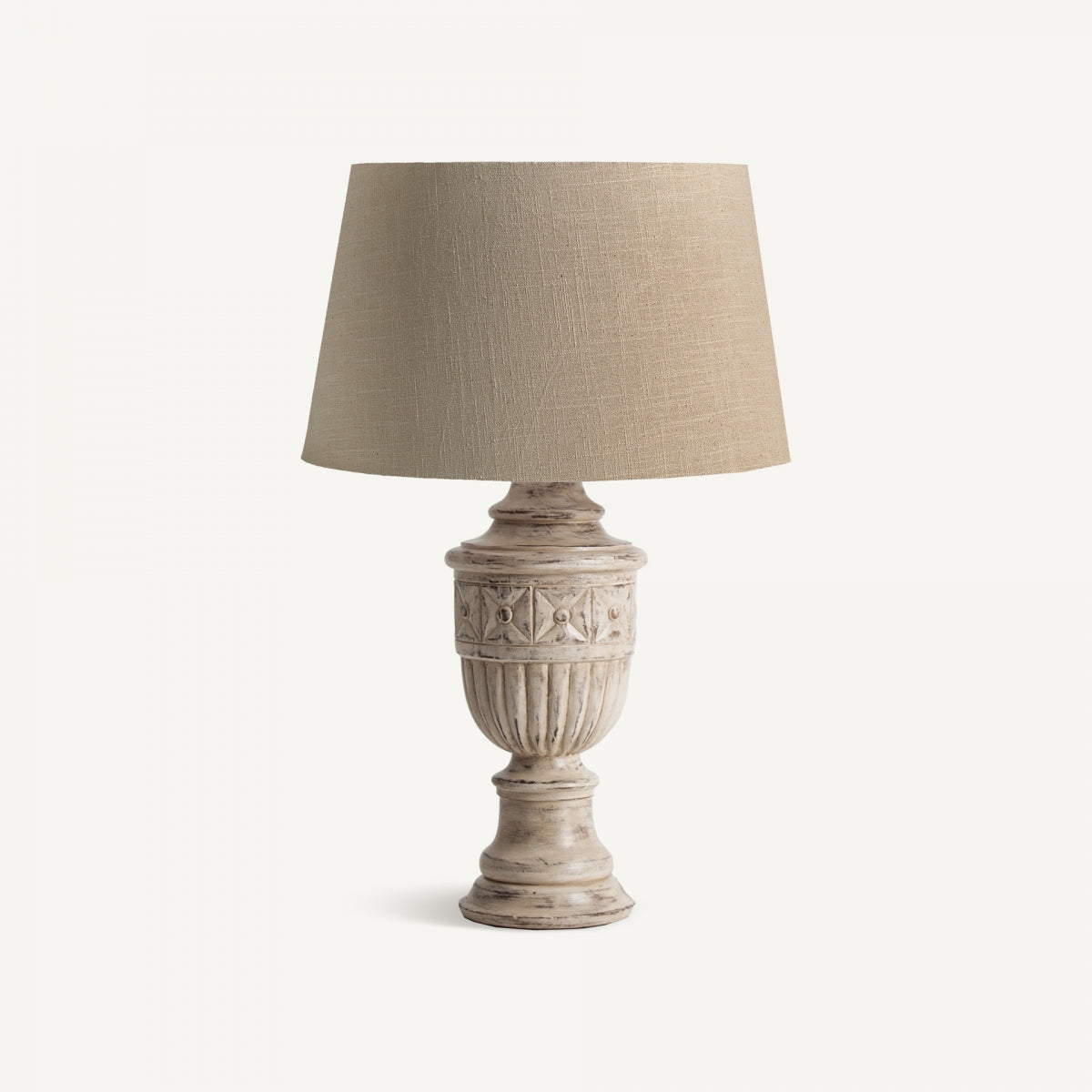 Una pantalla de tela beige reposa sobre la Lámpara de sobremesa en madera de mango Hout, que presenta una base rústica tallada, sobre un fondo blanco liso.