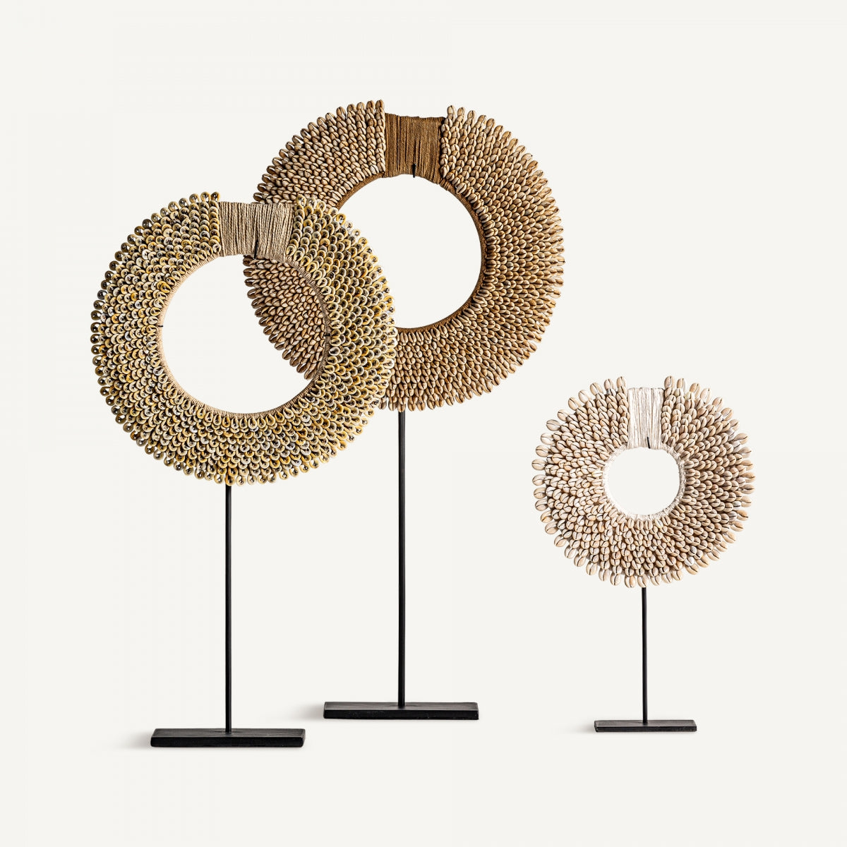 Tres piezas de "Figura decorativa en hierro y conchas Etnica", dos grandes en tono dorado y una pequeña en color beige, se apoyan sobre bases negras sobre un fondo liso, aportando un toque de estilo étnico y arte al hogar.