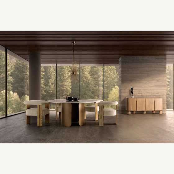Comedor moderno con una gran mesa de madera, seis sillas tapizadas, la lámpara colgante de hierro dorado Stowe A, ventanas de piso a techo y un aparador contra una pared texturizada con vista al bosque.