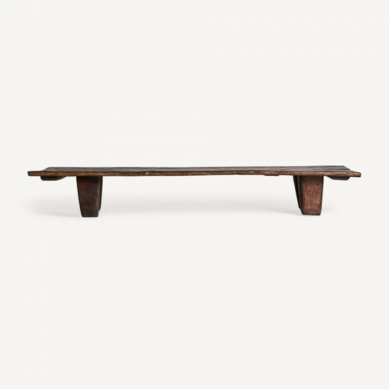 La mesa de centro rectangular en madera de teka Erara es un banco rústico y bajo de madera con un asiento alargado y patas gruesas y rectangulares hechas de teca maciza sobre un fondo liso.