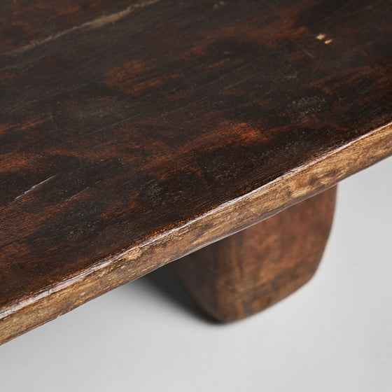 Primer plano de la mesa de centro rectangular de madera de teka Erara, con su esquina elaborada en teca maciza oscura. La superficie lisa y pulida y la veta visible realzan su diseño artesanal, elegantemente sostenido por una robusta pata.