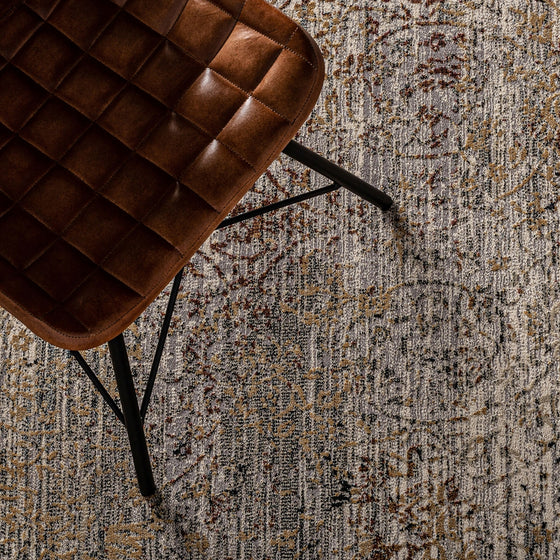 Una silla de cuero marrón con un cojín estampado cuadrado se asienta sobre la alfombra clásica en viscosa (300x200 cm) Ewa, en tonos grises, beige y marrones. La silla de la parte superior izquierda destaca la elegancia y confort del espacio.