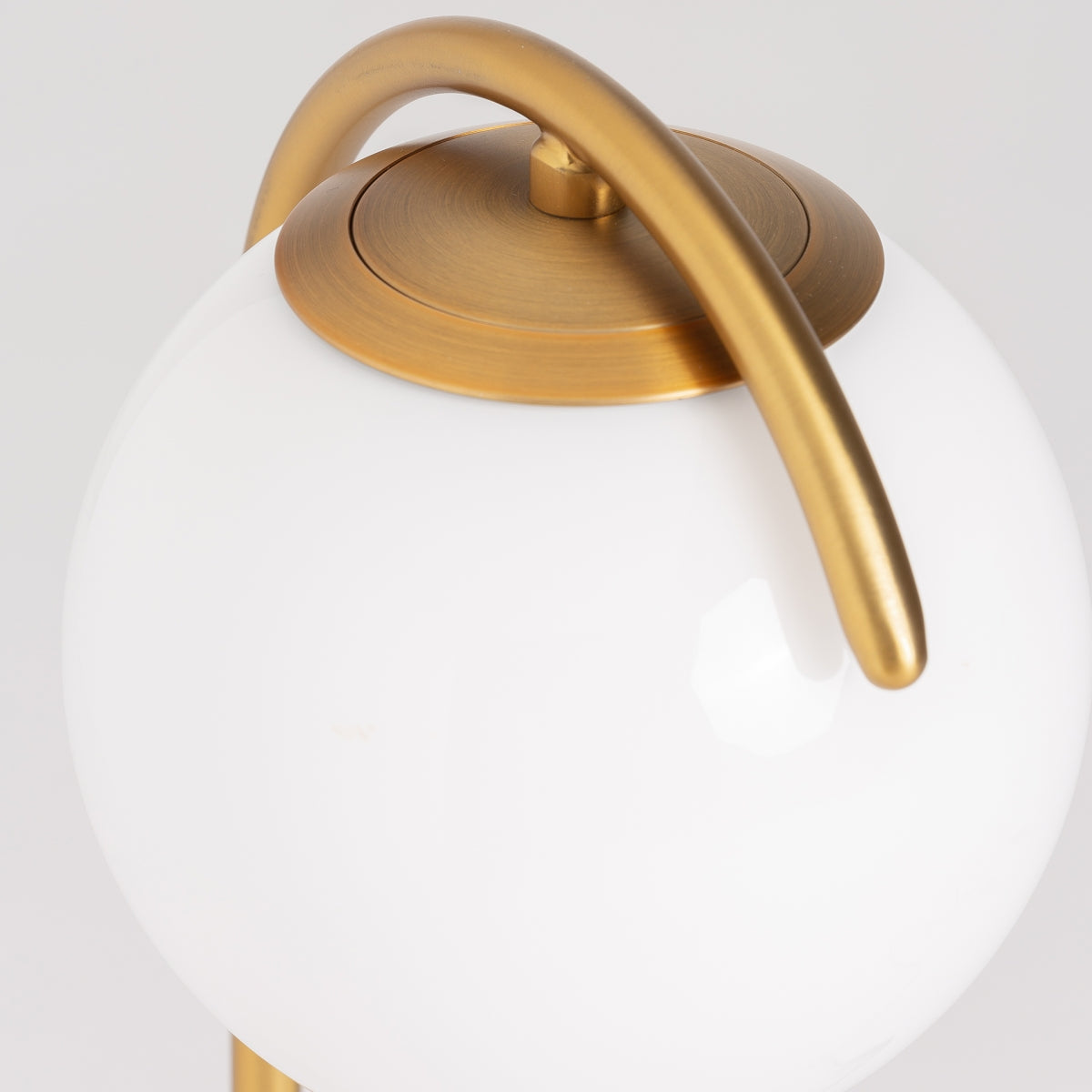 Un primer plano de la lámpara de mesa de hierro y mármol Holly, que presenta una pantalla esférica de vidrio blanco y un mango curvo de metal dorado, agrega sofisticación y lujo sobre un fondo liso.