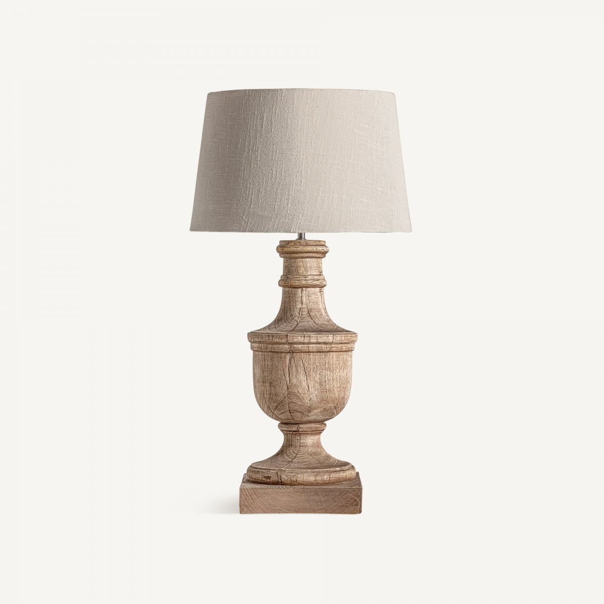 La lámpara de mesa en madera de mango Bolsa tiene una pantalla beige de lino y base tallada, con diseño provenzal, sobre fondo blanco.