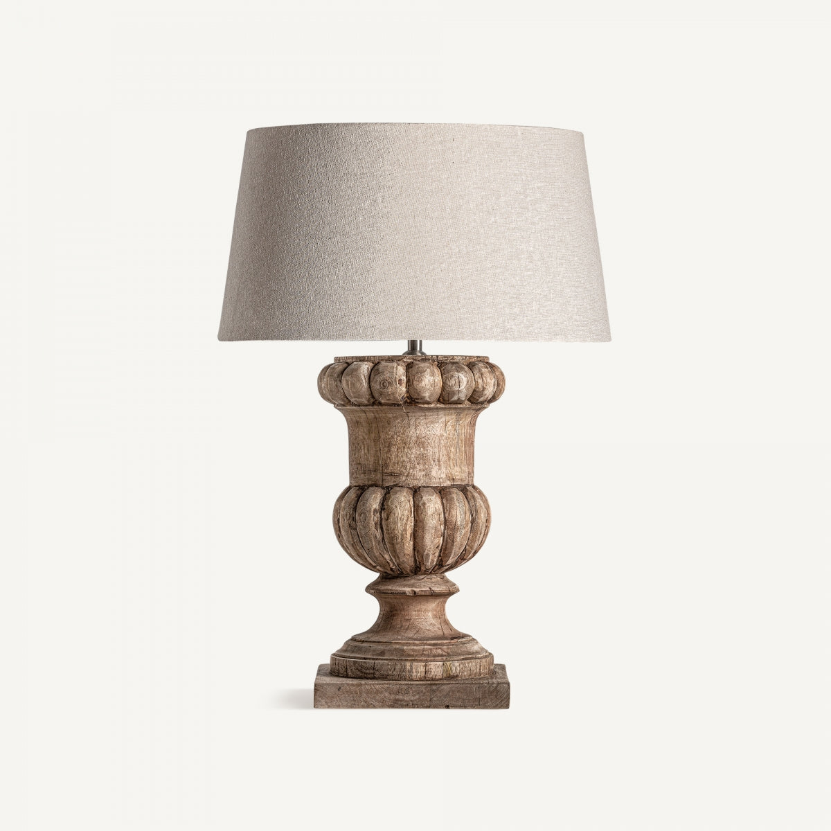 La Lámpara de sobremesa en madera de mango Nines presenta una pantalla beige texturizada y una base de madera tallada ornamentada, ideal para decoración rústica, que se muestra sobre un fondo blanco liso.