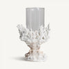 El Portavela en resina y cristal Arrecife S presenta un cuerpo de vidrio cilíndrico transparente elegantemente sostenido por una base en forma de coral blanco en elegante resina blanca, personificando el diseño moderno sobre un fondo liso.