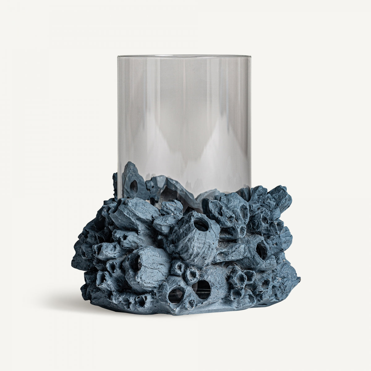 Un cilindro de vidrio transparente se posa sobre una base texturizada que se asemeja a formaciones de coral o rocas, capturando el diseño moderno del Portavela en resina azul Arrecife B.