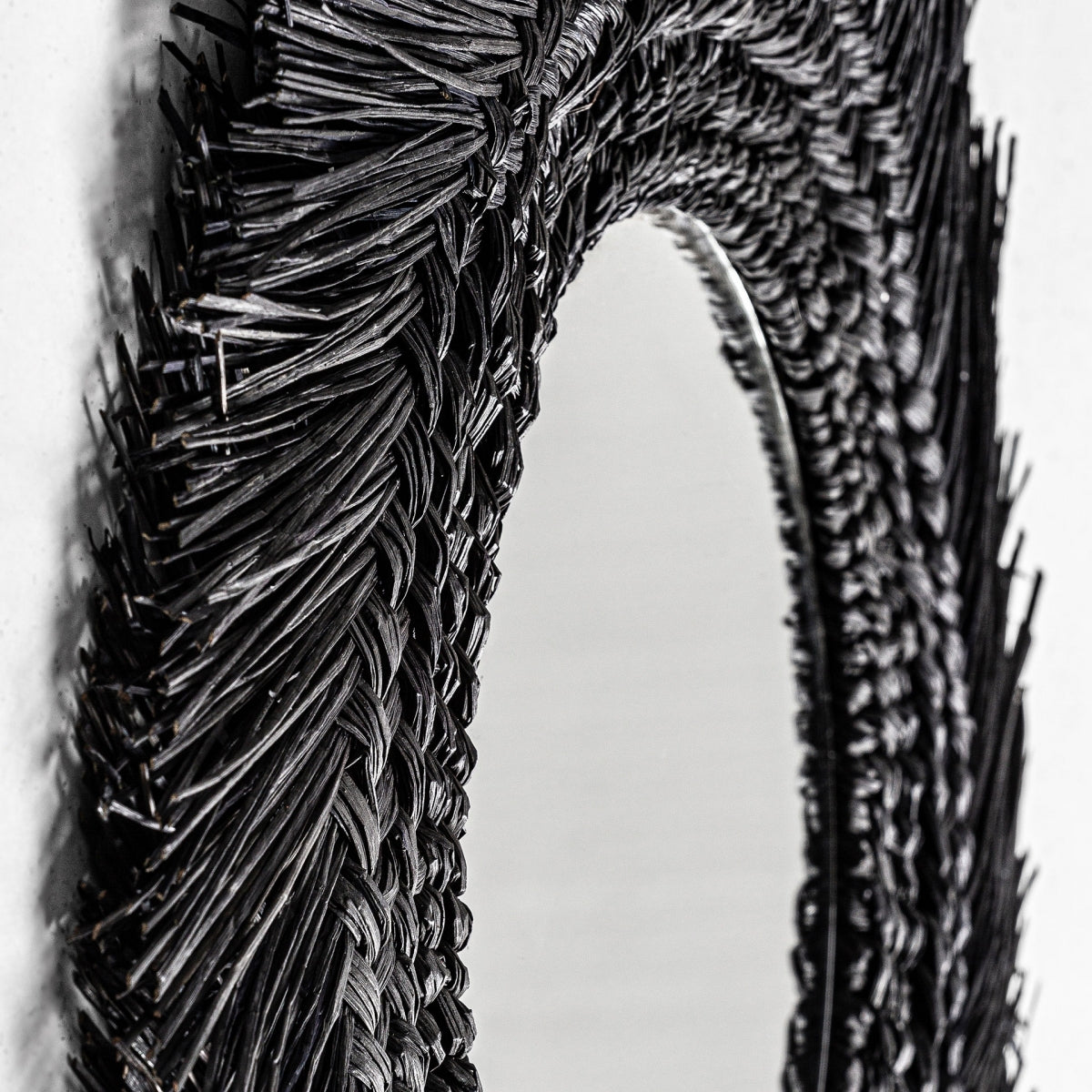 Primer plano del marco tejido negro del Espejo redondo en fibra natural (70x70 cm) de Desouk, que presenta un diseño contemporáneo texturizado y puntiagudo y una vista parcial del vidrio reflectante.