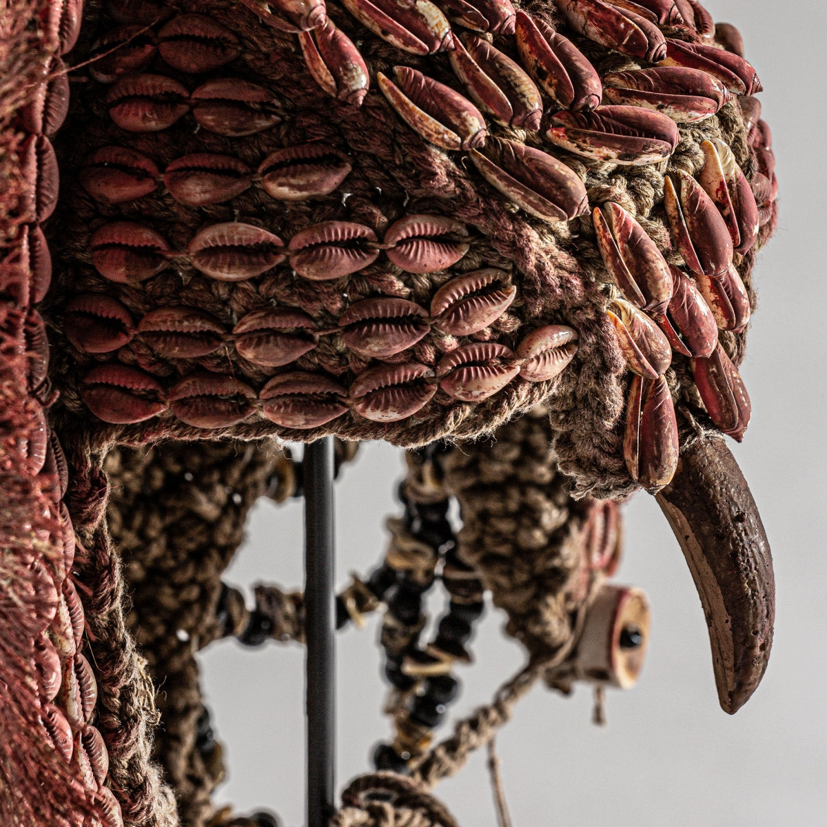 Primer plano de la figura decorativa en fibra natural étnica Kasai que muestra fibra natural texturizada, conchas de cauri rojas, cuentas negras y una garra curva prominente en un intrincado diseño étnico.