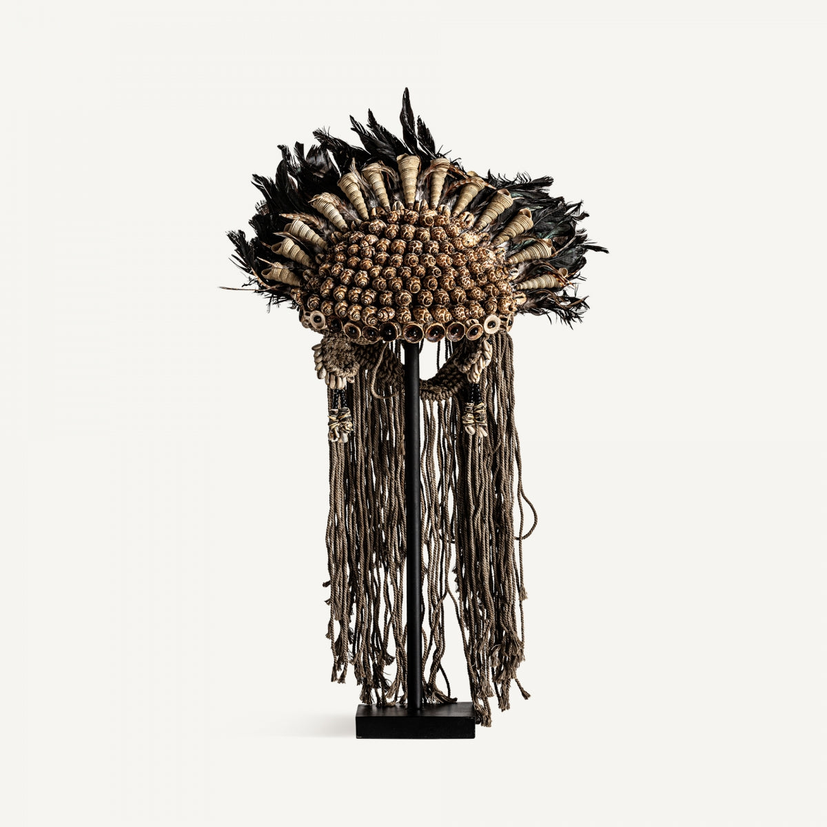 La figura decorativa en fibra étnica Azura presenta un diseño tribal con fibra natural, conchas y plumas negras, exhibida en soporte con largas borlas colgantes.
