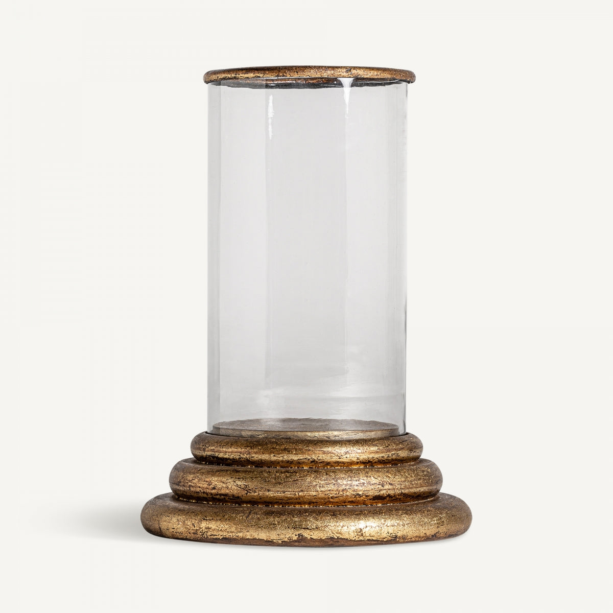 El Portavelas en madera y hierro mango presenta un cilindro de vidrio transparente con la parte superior abierta que descansa elegantemente sobre una base decorativa que muestra un diseño ornamental vintage y acabado en oro envejecido.
