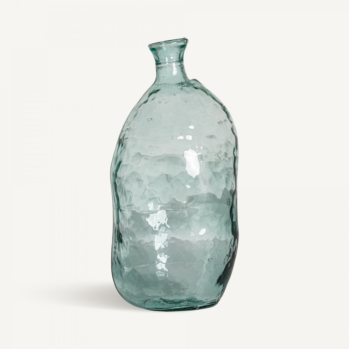 Un frasco de vidrio transparente y texturizado hecho de vidrio reciclado con una superficie irregular y un cuello inclinado: el Jarrón en vidrio reciclado Sumaya L, ideal para la decoración de interiores, que se muestra sobre un fondo claro y simple.