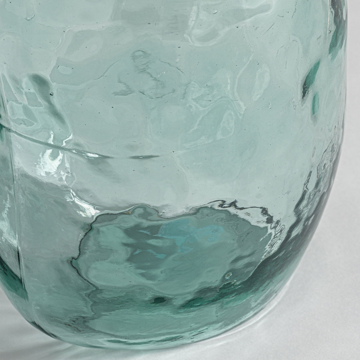 Primer plano de la mitad inferior del Jarrón de vidrio reciclado Sumaya L, con vidrio reciclado transparente y texturizado con un ligero tinte verde, perfecto para la decoración de interiores y exhibido sobre una superficie blanca.